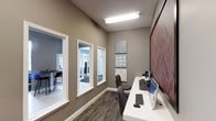 This single-floor space is designed with functionality and style in mind, offering a focused work environment that balances practicality with thoughtful aesthetics. The room features a sleek, elongated white desk equipped with a desktop computer, keyboard, and monitor, providing an efficient workstation setup. A cushioned ergonomic chair completes the workspace, ensuring comfort during extended use.

The walls are finished in a warm taupe-gray tone, lending a calm, professional atmosphere to the space. Overhead, a flush fluorescent light fixture provides consistent, even illumination throughout. Decorative wall art, featuring geometric and patterned tile-style prints in neutral and blue tones, adds visual interest without overwhelming the space.

A large-format display panel mounted on the adjacent wall suggests the room can also serve presentation or conferencing purposes. Interior windows with clean white trim line one wall, allowing natural light to filter in and offering sightlines into adjacent areas, which appear to include a communal or lounge space. The flooring consists of a patterned low-pile carpet in muted tones, reinforcing the professional character of the space.