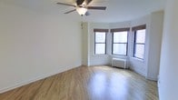 This single-floor urban residence offers a clean, functional layout with timeless architectural details. The main living area is an open, airy space finished with warm hardwood floors that reflect natural light, contributing to an inviting atmosphere. A classic ceiling fan is centrally mounted overhead, providing efficient air circulation throughout the room.

A standout feature is the bay window configuration set into a corner of the room, comprising multiple double-hung windows fitted with bamboo roller shades. This window arrangement allows generous natural light to fill the space while offering panoramic views of the surrounding cityscape. A traditional cast-iron radiator positioned beneath the windows provides reliable, even heating during colder months — a hallmark of classic urban residential architecture.

The walls are finished in a neutral white tone, providing a versatile backdrop that suits a variety of interior styles. Clean baseboards and well-maintained trim reflect careful upkeep throughout. The overall ambiance is bright, calm, and understated, with the city views and natural light serving as the primary focal points. This space presents a straightforward, practical living environment rooted in classic urban residential design.