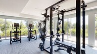 Located at 10945 Nuckols Road in Glen Allen, Virginia, this well-appointed fitness center offers a comprehensive workout environment across a single, thoughtfully organized floor. The space is divided into distinct workout zones, each designed to support a full range of exercise disciplines.

The cardio area is anchored by a row of treadmills, ellipticals, and a stationary spin bike, all positioned to take advantage of the floor-to-ceiling sliding glass windows and doors that line the perimeter. These expansive windows flood the space with natural light and provide unobstructed views of the surrounding greenery, creating an energizing atmosphere during workouts.

A dedicated strength training zone features a heavy-duty multi-functional rack equipped for squats, bench press, pull-ups, and cable exercises, complemented by a full rack of dumbbells. Adjacent to this is a functional training area with teal-toned carpet tiles, stability balls, TRX suspension trainers, medicine balls, a plyo box, and a wall-mounted flat-screen TV—ideal for group fitness or solo training sessions.

The interior design is clean and modern, with crisp white walls adorned with colorful artwork, recessed lighting, and a ceiling fan for added comfort. Light-toned linear carpet tiles throughout the main area and a dedicated sanitation station near the entrance reflect a well-maintained, user-focused facility.