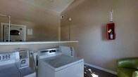 This single-floor laundry room is a dedicated utility space designed with practicality in mind. The room features commercial-grade Maytag top-load washing machines positioned along the wall, indicating a shared or multi-unit laundry facility. A sturdy wall-mounted shelf spans one side of the room, providing a convenient surface for sorting or folding laundry. The walls are finished in a warm neutral beige tone, and the space is equipped with standard electrical outlets and utility connections to support the appliances. A red fire safety panel is mounted on one wall, reflecting attention to building safety standards. The flooring appears to be a dark, durable material suited for a high-traffic utility environment. The angled ceiling adds a subtle architectural element to the otherwise straightforward space. Natural light filters into the room, casting soft patterns across the floor. Overall, the laundry room is a no-frills, functional space outfitted with the essential amenities needed for efficient laundry operations.