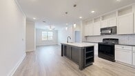 Located at 5025 Pilatus Way in Mebane, NC, this well-designed single-story home offers three bedrooms and three bathrooms within a thoughtfully laid-out floor plan. The open-concept kitchen and living area serves as the heart of the home, anchored by a striking two-tone island with a light stone countertop, integrated sink, and built-in shelving. White shaker cabinetry extends to the ceiling, complemented by a classic subway tile backsplash and black appliances, including a gas range, microwave, and refrigerator. Pendant lighting and recessed fixtures illuminate the space, while light wood-look flooring flows throughout the main living areas. The living room features a ceiling fan and a window that invites natural light. The primary bedroom is a comfortable retreat with carpeted flooring and access to a walk-in closet outfitted with wire shelving. Additional bedrooms also feature plush carpet, with one offering French doors that open to the outdoors. The bathrooms are finished with wood-look tile flooring, white soaking tubs, and dark-accented fixtures. A dedicated laundry room with wire shelving and utility hookups adds everyday convenience to this clean, move-in ready home.