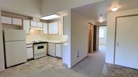 This single-floor space is designed with a practical, open layout that connects the kitchen to the rest of the unit through a central hallway. The kitchen is equipped with white cabinetry featuring wood-tone trim, a gas range with oven, a dishwasher, and a sink set beneath overhead cabinets. A fluorescent overhead light fixture illuminates the work area, and the kitchen floor is finished with neutral tile, providing a clean contrast to the adjoining spaces. A half-wall separates the kitchen from the carpeted hallway, which features a floor-level vent and leads toward additional rooms visible in the background. The hallway is lined with multiple doorways with matching wood-tone door frames, suggesting access to bedrooms and at least one bathroom. At the far end of the corridor, natural light filters in through a window, brightening the space and hinting at a more open room beyond. The flooring transitions from tile in the kitchen to carpet throughout the rest of the unit, contributing to a comfortable, residential feel. Neutral wall tones throughout create a clean and versatile backdrop suitable for a variety of interior styles.