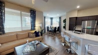 This single-floor residence showcases a thoughtfully designed open-concept layout that seamlessly connects the main living spaces. The living and dining area is anchored by warm hardwood-style flooring that extends throughout, creating a cohesive and inviting atmosphere. Multiple windows dressed with patterned navy drapes allow generous natural light to fill the space, enhancing its bright and airy feel. A ceiling fan overhead adds both function and comfort to the living area, which comfortably accommodates a full seating arrangement alongside a stylish coffee table. The dining area, positioned near a curved bay-style window arrangement, provides an elegant spot for gatherings and is well-suited for both casual and formal dining. Adjacent to the living space, a kitchen counter with bar seating offers a convenient transition between the cooking and living zones. The unit also features a private outdoor patio, enclosed by textured stucco walls and accessible through a teal-framed glass-panel door. The patio is furnished with a wicker seating set and a decorative area rug, creating a comfortable outdoor retreat with views of the surrounding landscaped grounds and parking area.