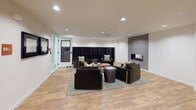 Located at 21212 International Blvd in SeaTac, Washington, this single-floor community amenity space is thoughtfully designed to support social interaction, recreation, and relaxation. The centerpiece of the space is a spacious lounge area finished with warm light wood flooring and crisp white walls. A tiled hexagon-patterned fireplace accent wall serves as a striking focal point, flanked by built-in dark bookshelves. Comfortable dark upholstered armchairs are arranged around a round drum coffee table atop a neutral area rug, creating an inviting conversational setting. Recessed lighting throughout ensures a well-lit, welcoming atmosphere. Adjacent to the lounge, a dedicated crafts room features teal carpeting, a central worktable with chairs, wall-mounted whiteboards, and ample overhead lighting — ideal for creative activities and group workshops. A corridor connects the various areas and is accented with framed artwork, including maps and decorative prints. A large wall-mounted Scrabble board adds a playful, interactive element to the hallway space. Further along, a game room area with a billiards table provides additional recreational options. Restroom facilities are conveniently accessible along the main corridor. The overall design balances modern aesthetics with functional, community-focused amenities.