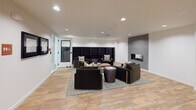 Located at 21212 International Blvd in SeaTac, Washington, this single-floor community amenity space is thoughtfully designed to support social interaction, recreation, and relaxation. The centerpiece of the space is a spacious lounge area finished with warm light wood flooring and crisp white walls. A tiled hexagon-patterned fireplace accent wall serves as a striking focal point, flanked by built-in dark bookshelves. Comfortable dark upholstered armchairs are arranged around a round drum coffee table atop a neutral area rug, creating an inviting conversational setting. Recessed lighting throughout ensures a well-lit, welcoming atmosphere. Adjacent to the lounge, a dedicated crafts room features teal carpeting, a central worktable with chairs, wall-mounted whiteboards, and ample overhead lighting — ideal for creative activities and group workshops. A corridor connects the various areas and is accented with framed artwork, including maps and decorative prints. A large wall-mounted Scrabble board adds a playful, interactive element to the hallway space. Further along, a game room area with a billiards table provides additional recreational options. Restroom facilities are conveniently accessible along the main corridor. The overall design balances modern aesthetics with functional, community-focused amenities.