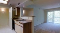 This single-floor residential space offers a practical and well-organized layout centered around an open-concept design. The kitchen is equipped with a double-basin sink, a dishwasher, and ample cabinetry featuring warm wood-toned upper cabinets complemented by white lower cabinets. A wraparound countertop with a peninsula-style overhang creates a natural divider between the kitchen and the adjacent living area, offering a convenient pass-through space ideal for casual dining or entertaining. The countertops carry a neutral, speckled finish that pairs well with the overall color palette. Overhead, HVAC vents are integrated into the ceiling, ensuring climate control throughout the space. The living area flows seamlessly from the kitchen, featuring soft carpeted flooring and light-toned walls that contribute to a bright and open atmosphere. The transition between the hard flooring of the kitchen and the carpeted living space is clearly defined, adding visual distinction between the two zones. The overall ambiance is clean, understated, and move-in ready, making it a highly functional space suited for everyday living.