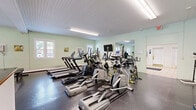 Located at 130 Clark St in Manchester, New Hampshire, this fitness facility offers a comprehensive exercise space designed to support a full range of workout routines. The single-floor layout is thoughtfully organized, beginning with a welcoming reception area finished with light tile flooring, a front desk counter with a wood-trimmed edge, and a small seating arrangement with leather chairs — ideal for check-ins and consultations.

The main exercise room is generously sized, with soft green walls and a white beadboard ceiling that lends the space a clean, bright aesthetic. Large windows along the walls allow natural light to fill the room, enhancing the open and airy atmosphere. Durable rubber tile flooring covers the workout area, providing both safety and comfort underfoot.

The cardio section is well-stocked with a row of treadmills, elliptical machines, stationary bikes, and cycling bikes — several equipped with individual digital consoles. Wall-mounted flat-screen televisions are positioned throughout for entertainment during workouts. A dedicated free-weight area features a full dumbbell rack alongside open floor space for stretching and functional training. A large mirror expands the visual depth of the room and aids in form monitoring. The facility also includes a specialty cable machine, a water cooler, and informational bulletin boards, making it a complete and functional fitness destination.