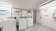 Located at 96 Isabella Street in Toronto's Church-Wellesley neighbourhood, this shared laundry facility offers a practical and well-equipped space for residents. The room is finished in crisp white walls and features a durable concrete floor, lending the space a clean, utilitarian aesthetic. Multiple coin-operated top-loading washing machines and front-loading dryers are arranged along one wall, providing ample capacity for resident use. The machines are serviced through a modern payment system, with instructional signage posted prominently for user convenience. Overhead fluorescent lighting ensures the room is well-lit throughout. The layout includes open floor space that allows for comfortable movement between machines. An accessible utility sink is available near the washers. The room is equipped with clearly marked exits, a fire alarm system, and a notice board for building communications, reflecting a commitment to safety and resident information. Exposed utility piping runs along the upper walls, consistent with the building's functional design approach. A separate storage or utility closet is accessible from within the laundry room. Overall, this facility is a straightforward, well-maintained communal amenity serving the building's residents.