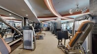 Located at 4108 Del Rey Ave in Marina del Rey, Los Angeles, this fitness center offers a comprehensive workout facility spread across a single, generously sized floor. The space is defined by its striking architectural design, highlighted by a curved, tiered ceiling with warm terracotta accents and integrated LED lighting that creates a dynamic and energizing ambiance throughout.

The facility is fully equipped with a wide array of professional-grade Cybex strength machines, including cable systems, overhead press stations, and functional training equipment. A dedicated row of cardio machines — including treadmills, ellipticals, and upright bikes — lines the perimeter wall, benefiting from large windows that invite natural light and offer views to the outdoors. Tile-clad structural columns punctuate the floor plan, adding an architectural character to the space.

A stretching and functional training area features padded floor mats and stability balls, providing space for warm-ups, cool-downs, and bodyweight exercises. Mounted flat-screen televisions are positioned throughout the facility for entertainment during workouts. Overhead fans ensure comfortable air circulation, while mirrored walls in select areas enhance the sense of openness. The neutral-toned carpet flooring and warm lighting combine to give the space a polished, welcoming atmosphere suited for a variety of fitness routines.
