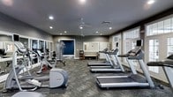 This fitness center offers a spacious, single-floor layout designed to accommodate a wide range of workout routines. The room is finished with neutral gray walls accented by a bold navy blue feature wall, giving the space a modern and energetic aesthetic. Recessed lighting throughout the ceiling ensures the room is well-lit, while two ceiling fans promote consistent air circulation during workouts.

The cardio section is prominently featured, housing a row of treadmills and elliptical machines positioned to make efficient use of the open floor plan. A multi-station cable and strength training unit is situated toward the back of the room, complemented by additional free-weight and resistance equipment. The carpeted flooring provides a practical surface suited for both cardio and strength training activities.

Large windows along one wall allow natural light to filter into the space, enhancing the overall atmosphere and creating a bright, inviting environment. A doorway connects to an adjacent room, suggesting additional amenities nearby. The layout is clean and organized, making it easy for users to navigate between equipment stations.