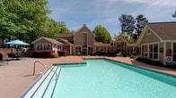 This outdoor pool area is a well-designed recreational amenity that combines functionality with a welcoming, resort-like atmosphere. At its centerpiece is a generously sized swimming pool with clear, turquoise water, accessible via stainless steel entry rails. The pool is framed by a broad concrete sundeck that provides ample space for lounging and socializing.

The deck is furnished with multiple slatted metal chaise lounges and shaded seating areas featuring umbrellas, offering residents comfortable options for relaxation throughout the day. Surrounding the pool area, vibrant landscaping includes manicured shrubs, ornamental red-leafed plants, and mature deciduous trees that provide natural shade and a sense of privacy.

Adjacent to the pool is a clubhouse-style building with a gabled roofline, taupe lap siding, and large glass double doors, providing convenient access to interior facilities. A covered pergola structure on one side of the deck adds architectural interest and additional shaded seating. The pool area is enclosed by black metal fencing, ensuring a secure and defined space. The combination of lush greenery, thoughtful seating arrangements, and well-maintained facilities makes this pool area an inviting outdoor retreat.