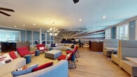 Located at 1560 San Luis Rd in Tallahassee, Florida, this single-floor collaborative workspace is designed to inspire productivity and creativity. The centerpiece of the space is a generously sized open lounge area anchored by a curved sectional sofa, bold red accent chairs, and a large ottoman, all arranged on light hardwood flooring. Cylindrical side stools in teal and gray add playful pops of color throughout. A striking sputnik-style chandelier hangs overhead, complemented by ceiling fans and recessed lighting, creating a bright and welcoming atmosphere. The focal wall features a dramatic starburst mural with words such as "Solutions," "Reliability," "Satisfaction," and "Efficiency" radiating outward in bold typography, reinforcing the space's professional and motivational character. To one side, glass-partitioned conference rooms with white framing offer transparent, collaborative meeting areas furnished with wooden tables and blue rolling chairs. A dedicated computer workstation area provides desktop setups along the back wall, ideal for focused work. Large windows along the perimeter allow generous natural light and offer views of the surrounding greenery. The overall design blends a modern, energetic aesthetic with functional, multi-use zones suited for group collaboration, meetings, and individual work.