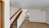 Located at 1602 Valparaiso St in Valparaiso, Indiana, this two-story unit offers a straightforward, functional layout spread across two levels. The main level features an open living area finished with wall-to-wall carpet, crisp white walls, and warm wood baseboards that carry consistently throughout the space. A large front window allows natural light to fill the room, complemented by a tiled entryway near the front door. The kitchen is efficiently designed in an L-shaped configuration, featuring white cabinetry with wood-trimmed accents, ample upper and lower storage, a double-basin sink, a gas range, dishwasher, and tile flooring. A staircase with a warm wood handrail connects the main level to the upper floor. The upper level contains multiple carpeted bedrooms, each with large windows fitted with blinds that provide natural light and outdoor views. The bathroom includes a full tub-and-shower combination with a chrome curtain rod, a toilet, and tile flooring. Throughout both floors, the design maintains a clean, neutral palette that provides a versatile backdrop adaptable to a variety of furnishing styles.