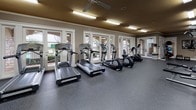 This fitness center is a spacious, single-floor facility designed to support a full range of workout routines. The layout is thoughtfully organized into distinct zones, creating an efficient and comfortable environment for exercise. The cardio area is lined with treadmills, ellipticals, and stationary bikes, all positioned along a wall of large windows and glass doors that flood the space with natural light and offer views of the outdoor surroundings. A ceiling fan overhead enhances airflow in this section of the facility. The strength training area features multiple cable and weight machines, along with a dedicated free weights station equipped with a tiered dumbbell rack, an adjustable bench, and a wall-mounted television for entertainment during workouts. A standout architectural detail is the natural stacked stone accent wall, which adds warmth and texture to the space and is complemented by a decorative wall clock. Floor-to-ceiling mirrors on one side of the room visually expand the space and allow users to monitor their form. The entire facility is finished with durable, speckled rubber flooring, and overhead fluorescent lighting ensures consistent illumination throughout. Crown molding along the upper walls and neutral-toned walls with warm ceiling finishes contribute to a polished, inviting ambiance.
