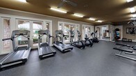 This fitness center is a spacious, single-floor facility designed to support a full range of workout routines. The layout is thoughtfully organized into distinct zones, creating an efficient and comfortable environment for exercise. The cardio area is lined with treadmills, ellipticals, and stationary bikes, all positioned along a wall of large windows and glass doors that flood the space with natural light and offer views of the outdoor surroundings. A ceiling fan overhead enhances airflow in this section of the facility. The strength training area features multiple cable and weight machines, along with a dedicated free weights station equipped with a tiered dumbbell rack, an adjustable bench, and a wall-mounted television for entertainment during workouts. A standout architectural detail is the natural stacked stone accent wall, which adds warmth and texture to the space and is complemented by a decorative wall clock. Floor-to-ceiling mirrors on one side of the room visually expand the space and allow users to monitor their form. The entire facility is finished with durable, speckled rubber flooring, and overhead fluorescent lighting ensures consistent illumination throughout. Crown molding along the upper walls and neutral-toned walls with warm ceiling finishes contribute to a polished, inviting ambiance.