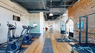 Located at 698 Martin Luther King Jr Blvd in Macon, GA, this fitness center combines industrial character with modern functionality in a single, thoughtfully designed floor. The space features warm hardwood flooring throughout, exposed brick accent walls, and soaring ceilings with visible ductwork and structural beams that lend an authentic urban aesthetic. Pendant lighting fixtures hang at varying heights, providing ample illumination across the workout area.

The exercise room is well-appointed with a comprehensive selection of equipment, including treadmills, elliptical trainers, a stationary bike, a cable machine, free weights, and weight plates — catering to both cardio and strength training needs. Rubber exercise mats are strategically placed to define workout zones. A wall-mounted television keeps users entertained during workouts, while large multi-pane windows along one wall flood the space with natural light and offer views of the surrounding neighborhood.

A standout arched glass-and-steel door serves as a striking architectural feature, connecting the workout area to a bright, open lounge zone. This adjoining area includes a concrete ping-pong table, vibrant red armchairs, and curated wall art, providing a social and recreational counterpoint to the active fitness space. The overall ambiance is energetic yet refined, blending historic building character with contemporary amenities.