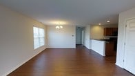 This single-floor space offers an open-concept layout that seamlessly connects the living area to the kitchen. The living space is anchored by warm, dark hardwood flooring that extends throughout, creating a cohesive and inviting atmosphere. A classic chandelier serves as a focal point overhead, adding a touch of elegance to the room. Natural light filters in through a double-pane window fitted with blinds, brightening the neutral white walls and contributing to an airy feel. Recessed lighting provides additional illumination in key areas. The kitchen is partially separated by a raised counter with dark countertops, offering a functional breakfast bar-style divide. Wood-toned upper cabinetry and a visible sink add practicality to the space. A doorway visible in the background leads to additional areas of the unit, suggesting a thoughtful floor plan. The clean, simple design with neutral tones throughout creates a versatile canvas well-suited for a variety of interior styles.