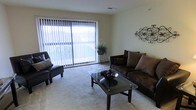 This single-floor space offers a clean, functional layout designed for comfortable everyday living. The living area serves as the centerpiece of the home, highlighted by a large sliding glass door dressed with vertical blinds that allow natural light to filter throughout the room while providing a view of the outdoors. The soft, plush wall-to-wall carpeting adds warmth underfoot and contributes to the overall cozy ambiance of the space. Neutral-toned walls create a versatile backdrop, complemented by decorative wall accents that add a touch of character. The living area comfortably accommodates a full seating arrangement, including a sofa and accent chair paired with a glass-top coffee table, demonstrating the generous and practical use of the available space. A decorative plant stand near the window introduces a natural element, enhancing the light and airy atmosphere. Ceiling-mounted vents indicate a central climate control system for year-round comfort. Overall, this space balances practicality with an inviting aesthetic, making it a comfortable and functional home environment.