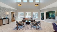 Located at 3310 W 84th St in Inglewood, California, this single-story community clubhouse offers a thoughtfully designed gathering space that blends modern aesthetics with functional comfort. The centerpiece of the facility is a dramatic main lounge, defined by soaring double-height ceilings, oversized windows, and a row of statement pendant lights with a geometric honeycomb design. The open layout is anchored by a patterned terrazzo-style floor, with a curated seating arrangement of leather and wood-framed chairs surrounding a resin-top coffee table, all set atop a layered blue area rug. Large windows along the rear wall frame views of the outdoor pool area, flooding the space with natural light. A secondary lounge features vaulted ceilings, warm hardwood flooring, large sliding glass doors that open to an outdoor patio, and contemporary abstract artwork. This room is furnished with deep navy blue sofas and woven accent chairs, creating an inviting, casual atmosphere. A well-appointed kitchenette serves the facility with white shaker cabinetry, a beverage cooler, microwave, coffee station, and a water dispenser. The front entry area includes a reception counter and additional seating, completing this versatile and welcoming community space.