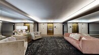 This sophisticated elevator lobby delivers a refined, hotel-inspired ambiance through a carefully curated blend of luxurious materials and elegant design. The space is anchored by warm hardwood flooring that transitions seamlessly beneath a large, dark patterned area rug, lending depth and visual contrast to the interior. Polished stone wall cladding in soft cream tones lines the perimeter, complemented by gleaming brass elevator doors that serve as striking architectural focal points. Full-length mirrored wall panels extend the perceived scale of the space, reflecting the interior and amplifying the ambient glow of recessed cove lighting integrated into the ceiling. The smooth, tray-style ceiling with its subtle illumination creates a calm, welcoming atmosphere. Furnishings are tastefully arranged throughout, including a blush pink velvet sofa, neutral upholstered armchairs, and a decorative gold console table adorned with soft floral accents. Dark wood-framed glass entry doors provide a stately transition point, reinforcing the classic yet contemporary character of the space. Together, these elements create a polished, welcoming environment that balances timeless elegance with modern sensibility.
