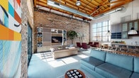 Located at 695 S Santa Fe Ave in the heart of Los Angeles' Arts District, this single-floor space masterfully blends industrial heritage with modern design. Expansive exposed brick walls stretch from floor to ceiling, complemented by striking wood-beam and timber-truss ceilings with visible ductwork that add an authentic warehouse character throughout.

The open living area is generously proportioned, bathed in natural light streaming through oversized steel-frame windows. Polished concrete floors anchor the space, while carefully curated furnishings — including plush sofas, accent armchairs, and layered rugs — create a warm, inviting atmosphere against the raw architectural backdrop. A built-in media and shelving unit serves as a functional focal point along the brick wall.

The kitchen is a standout feature, boasting a substantial stainless steel island with bar seating, dark wood cabinetry with glass-front uppers, white subway tile backsplash, and dramatic industrial pendant lighting. The kitchen opens seamlessly to the living area, reinforcing the loft's open-plan flow.

Large multi-pane windows and glass doors throughout the space frame urban views and draw in abundant daylight, connecting the interior to its dynamic surroundings. The overall ambiance is sophisticated yet approachable — a refined industrial aesthetic ideal for both living and entertaining.