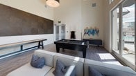 This contemporary game room is designed for both entertainment and relaxation, offering a well-appointed space with an impressive array of recreational amenities. The room's most striking architectural feature is its soaring ceiling height, which creates an open, airy atmosphere throughout. Large floor-to-ceiling windows along one wall flood the space with abundant natural light while providing outdoor views, further enhancing the sense of openness.

The game room is anchored by a sleek, dark-finished pool table positioned prominently at the center, complemented by a wooden shuffleboard table along one wall. A wall-mounted flat-screen television provides additional entertainment options. Decorative elements include a bold geometric textured accent wall panel and colorful abstract wall art, adding visual interest against the clean white walls.

The lounge area features a comfortable sectional sofa, accent armchairs, and round coffee tables arranged atop a richly patterned area rug, creating an inviting space for socializing. Light wood-toned flooring runs throughout, tying the space together cohesively. The overall design balances modern aesthetics with functional recreational use, making it an ideal gathering space.