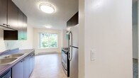 Located at 1820 NE 104th Ave in Portland, Oregon, this two-bedroom, one-bathroom unit offers a practical and well-organized single-floor layout. The living area features warm wood-look flooring and clean white walls, creating a bright and open feel throughout the main space. The galley-style kitchen is a standout area, equipped with dark espresso cabinetry, distinctive purple countertops, a stainless steel range, refrigerator, and a sink with ample counter space on both sides. A window at the end of the kitchen fills the space with natural light and a view of surrounding greenery. Both bedrooms are finished with neutral carpet and feature sliding double-door closets for convenient storage. Each bedroom includes a baseboard heating unit and windows that allow in natural light with views of the neighborhood. The bathroom is cleanly appointed with wood-look tile flooring, a single vanity with dark cabinetry, and a full soaking tub with shower, complemented by grab bars and bright white surroundings. The overall design is clean and functional, with a consistent neutral palette throughout that offers a versatile canvas for personalization.