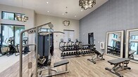 Located at 3260 Stockton Street in North Fort Myers, FL, this fitness center offers a comprehensive and thoughtfully designed exercise facility spread across a single, open floor. The main exercise room impresses with soaring ceilings and an abundance of natural light streaming through large, floor-to-ceiling windows that offer views of the surrounding outdoor landscape. Stylish geometric chandelier pendant lights add an upscale touch to the space, complementing the warm, wood-look plank flooring throughout.

The facility is fully equipped with a wide array of strength and cardio equipment, including cable machines, a multi-rack dumbbell station with adjustable bench, treadmills with personal display screens, stair climbers, and ellipticals. A dedicated studio room features bold, colorful wall art, Matrix cycling bikes, and a weight bench with barbell rack, making it ideal for focused training sessions.

A separate flex room with textured accent wallpaper and full-length wall mirrors provides an open area for stretching, yoga, or group exercise. A built-in media console with a mounted flat-screen TV, integrated water fountain, and storage cabinetry serves as a convenient amenity station. The overall ambiance is modern and polished, offering both functionality and an elevated aesthetic for an engaging workout environment.