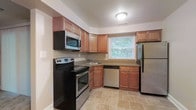 This single-story space includes a functional kitchen designed for efficient use. The kitchen features warm-toned wood cabinetry that lines the walls, providing ample upper and lower storage throughout. Dark laminate countertops offer generous prep space, complementing the stainless steel appliance suite, which includes an electric range with oven, an over-the-range microwave, a dishwasher, and a full-size refrigerator. A single-basin sink is positioned beneath a window that allows natural light to fill the space and offers a view of outdoor greenery. The light beige tile flooring adds a neutral, clean aesthetic that ties the space together. The layout follows a practical L-shaped configuration, maximizing workflow between cooking and prep areas. Recessed flush-mount ceiling lighting ensures consistent illumination throughout. The neutral wall tones and straightforward design create a clean, understated ambiance suited for everyday use.