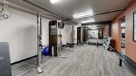 Located at 304 W North St in Waukesha, Wisconsin, this fitness center offers a comprehensive exercise environment within a single, efficiently organized floor. The space is divided into distinct workout zones, making it easy to navigate between cardio and strength training areas. Along one wall, a row of Precor treadmills and elliptical machines—each equipped with individual monitors—provides a dedicated cardio section, with large mirrors enhancing the sense of depth. The strength training area features multiple cable machines with integrated display screens, adjustable benches, and a barbell/weight plate station for functional and compound movements. A free weight rack stocked with a range of dumbbells rounds out the resistance training options, while stability balls add versatility for core and flexibility work. A seated lower-body machine and additional functional training equipment round out the offerings. The room is finished with durable, patterned carpet flooring and illuminated throughout by overhead fluorescent lighting. Exposed overhead piping and a neutral color palette accented by bold terracotta-orange wall panels give the space an energetic, no-frills aesthetic. Storage is provided by a multi-drawer filing cabinet, and a sanitation station ensures a clean environment. The space is accessible via a dedicated entry door.
