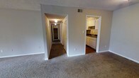 This single-floor unit is thoughtfully laid out to maximize functionality and flow. The main living area is generously sized, featuring neutral wall-to-wall carpeting that lends a comfortable, understated feel underfoot. A textured popcorn ceiling runs throughout, adding a classic residential character to the space. The focal point of the living area is a large sliding glass door that fills the room with natural light and opens to a private balcony with a metal railing, offering views of surrounding greenery.

From the living area, the layout branches into additional spaces. A doorway to one side opens directly into the kitchen, which features white cabinetry, a dishwasher, under-cabinet range hood, and dark countertops. The kitchen is compact yet well-equipped, with tile flooring that contrasts the carpeted living spaces.

A hallway extends further into the unit, with arched and standard doorways leading to what appear to be additional rooms and closet storage with louvered doors. Overhead lighting in the hallway provides practical illumination. The overall ambiance is clean and neutral, offering a versatile canvas ready for personalization.
