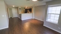 This single-floor space is designed with an open-concept layout that seamlessly connects the living area and kitchen, creating a cohesive and functional environment. The dark hardwood-style flooring runs throughout, providing a rich, grounding contrast to the light gray walls and crisp white baseboards.

The kitchen is positioned centrally and defined by a raised peninsula with a light-toned countertop, offering both workspace and a casual dining or gathering surface. Dark cabinetry overhead and below adds depth, while stainless steel appliances — including a refrigerator and microwave — bring a modern, practical finish. Track lighting above the kitchen ensures well-lit food preparation areas.

The expansive living area benefits from an abundance of natural light entering through a window on the right side of the space. A ceiling fan is centrally mounted, adding to the room's comfort and airflow. Recessed lighting complements the track fixtures, ensuring even illumination throughout.

The overall ambiance is clean, modern, and welcoming, with a neutral palette that allows for versatile furnishing and personalization.