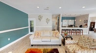 This well-appointed single-floor space offers an open-concept layout that seamlessly connects the living, dining, and kitchen areas, creating a cohesive and inviting environment. The living area is anchored by comfortable seating arrangements, including a cream sofa and accent chairs, all set atop light wood-style flooring. Bold teal accent walls, trimmed with a crisp white chair rail, add a striking pop of color and visual interest throughout the space, complementing the neutral warm-beige tones on adjacent walls. Decorative wall art and vibrant area rugs contribute to the warm, stylish ambiance.

The kitchen is visible from the living area and features a raised bar counter with wood-topped barstools, pendant lighting, and modern appliances, making it ideal for both casual gatherings and entertaining. A separate dining area is bathed in natural light, courtesy of large wraparound windows that offer views of the lush outdoor greenery and surrounding grounds. The dining space accommodates a full-sized table with colorful mixed seating, lending an eclectic yet cohesive aesthetic.

Recessed lighting throughout ensures the space remains bright and well-lit, while access doors lead to additional rooms and the exterior. Overall, this space strikes a balance between functional design and cheerful, contemporary décor.