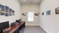 This single-floor space functions as a dedicated computer or business center room. The room features neutral, light-colored walls that create a clean and focused atmosphere, complemented by gray carpeting underfoot that adds a degree of comfort and sound absorption. A wooden workstation desk runs along one wall, equipped with dual monitors, a keyboard, and a printer, making it well-suited for productivity tasks. Black task chairs are positioned at the desk, providing functional seating for multiple users. A centrally placed window allows natural light to filter into the room, brightening the otherwise understated interior. The window overlooks a lightly wooded exterior view, adding a subtle connection to the outdoors. Decorative pennants and small framed pieces are mounted on the walls, lending the space a casual, personalized character. The ceiling features a standard HVAC vent, ensuring climate comfort throughout the room. Overall, the space is straightforward and efficiently arranged, prioritizing function while maintaining a tidy, welcoming environment suitable for work or study purposes.