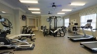 This fitness center offers a comprehensive workout environment designed to accommodate a variety of exercise routines. The space is finished with durable speckled rubber flooring, ideal for heavy equipment and high-traffic use, while crown molding along the ceiling adds a refined architectural touch. Overhead, multiple flush-mount fluorescent light fixtures and two ceiling fans ensure the room remains bright and well-ventilated throughout.

The facility is outfitted with an impressive selection of equipment. A multi-station cable and weight machine serves as the centerpiece, surrounded by free-weight benches, a dumbbell rack, and an assortment of cardio machines including treadmills, a stationary bike, and an elliptical. A wall-mounted flat-screen television provides entertainment during workouts, while natural light filters in through a window dressed with a Roman shade.

The layout is open and thoughtfully arranged, allowing users to move freely between stations without congestion. A doorway leads to an adjacent room, suggesting additional amenities or utility space nearby. Overall, the fitness center combines functionality with a clean, polished aesthetic, making it a practical and inviting space for daily exercise.