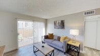 This well-appointed single-floor residence offers a clean, modern aesthetic with a welcoming living area at its heart. The living room is finished with light wood-style flooring and a textured ceiling, creating a warm yet contemporary feel. A large sliding glass door dominates one wall, fitted with vertical blinds that allow for flexible light control, and opens directly onto an outdoor balcony — flooding the interior with natural light and providing a pleasant connection to the outdoors. The living space comfortably accommodates a plush grey sofa accented with mustard and multicolored throw pillows, paired with a minimalist wood-and-metal coffee table and a coordinating side table. Wall décor adds a touch of urban personality to the neutral, light grey walls. The overall ambiance is bright, clean, and inviting, with a cohesive color palette of soft greys, warm yellows, and natural wood tones that lend the space a casual yet polished character. The layout is thoughtfully arranged to maximize the sense of openness, making the living area feel airy and functional — ideal for both everyday living and relaxed entertaining.