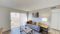 This well-appointed single-floor residence offers a clean, modern aesthetic with a welcoming living area at its heart. The living room is finished with light wood-style flooring and a textured ceiling, creating a warm yet contemporary feel. A large sliding glass door dominates one wall, fitted with vertical blinds that allow for flexible light control, and opens directly onto an outdoor balcony — flooding the interior with natural light and providing a pleasant connection to the outdoors. The living space comfortably accommodates a plush grey sofa accented with mustard and multicolored throw pillows, paired with a minimalist wood-and-metal coffee table and a coordinating side table. Wall décor adds a touch of urban personality to the neutral, light grey walls. The overall ambiance is bright, clean, and inviting, with a cohesive color palette of soft greys, warm yellows, and natural wood tones that lend the space a casual yet polished character. The layout is thoughtfully arranged to maximize the sense of openness, making the living area feel airy and functional — ideal for both everyday living and relaxed entertaining.