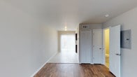 This two-floor space is designed with a clean, neutral aesthetic that creates a bright and functional living environment. The main living area showcases rich, dark hardwood-style flooring that contrasts beautifully with the crisp white walls and baseboards, lending the space a polished, contemporary feel. A flush-mounted ceiling light provides overhead illumination, while a sliding glass door dressed with vertical blinds allows an abundance of natural light to filter into the room, seamlessly connecting the interior to the outdoors.

A transition to light-toned tile flooring marks the entry into an adjoining area near the sliding door, suggesting a defined transitional zone between the living space and an outdoor access point. To one side, a set of louvered bi-fold closet doors offers practical storage, complemented by a standard interior door that leads to an additional room, visible with warm lighting. An overhead HVAC vent is also present, indicating a central climate control system throughout the unit. The overall ambiance is light, airy, and move-in ready, with a straightforward layout that maximizes usability across both floors.