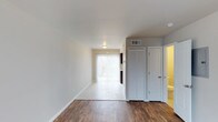 This two-floor space is designed with a clean, neutral aesthetic that creates a bright and functional living environment. The main living area showcases rich, dark hardwood-style flooring that contrasts beautifully with the crisp white walls and baseboards, lending the space a polished, contemporary feel. A flush-mounted ceiling light provides overhead illumination, while a sliding glass door dressed with vertical blinds allows an abundance of natural light to filter into the room, seamlessly connecting the interior to the outdoors.

A transition to light-toned tile flooring marks the entry into an adjoining area near the sliding door, suggesting a defined transitional zone between the living space and an outdoor access point. To one side, a set of louvered bi-fold closet doors offers practical storage, complemented by a standard interior door that leads to an additional room, visible with warm lighting. An overhead HVAC vent is also present, indicating a central climate control system throughout the unit. The overall ambiance is light, airy, and move-in ready, with a straightforward layout that maximizes usability across both floors.
