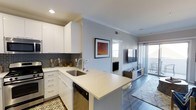 This single-floor residence showcases a thoughtfully designed open-concept layout that connects the kitchen and living areas into a cohesive, flowing space. The modern kitchen is outfitted with crisp white cabinetry, sleek quartz countertops, and a classic subway tile backsplash in a charcoal hue that adds depth and contrast. A peninsula with bar seating extends from the main counter, providing additional workspace and casual dining options. Stainless steel appliances, including a built-in dishwasher, complement the clean, contemporary aesthetic. The kitchen opens directly into the living area, which features warm hardwood-style flooring, crown molding, and a wall-mounted flat-screen television set above a low-profile media console. Natural light filters in through a large window and sliding glass doors that lead to an outdoor balcony or patio, enhancing the airy ambiance of the space. A separate room visible beyond the living area offers additional functionality, whether as a bedroom or dedicated workspace. The neutral color palette of soft grays and whites throughout creates a calm, modern atmosphere that feels both welcoming and refined.