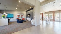 This vibrant, single-floor community clubhouse is thoughtfully designed to offer a dynamic and welcoming atmosphere for socializing and relaxation. The spacious main gathering area features large-format herringbone-patterned tile flooring, soaring ceilings with recessed lighting, and an abundance of natural light streaming in through floor-to-ceiling glass doors that open toward an outdoor area. The open layout flows seamlessly between zones, each with its own distinct character.

The lounge area is anchored by a bold slate-blue accent wall, a wall-mounted flat-screen TV, and a sleek linear electric fireplace. Sculptural, artistic furniture — including a white hand-shaped chair and an organically-patterned media console — add playful, contemporary flair. A patterned carpet with coral accents defines the seating zone, which includes a navy sofa and vibrant red accent chairs.

Adjacent to the lounge is a game room highlighted by a striking red abstract wall mural, a billiards table, and bar-height seating — creating an energetic entertainment hub. A cozy dining nook features a round table, white modern chairs, a globe chandelier, and bold red drapery that frames views of lush greenery outside.

A built-in cabinetry station with dark wood-grain drawers, white countertops, and curated décor accents provides a polished service or workspace area. Throughout the space, contemporary artwork and thoughtful styling reinforce a cohesive, upscale aesthetic.