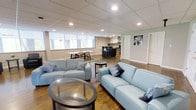 This well-appointed single-floor space offers an open-concept layout that seamlessly connects a generous living area, a dining zone, and a sleek kitchenette. The living area is anchored by two light blue sofas arranged around a round glass-top coffee table, creating a welcoming and cohesive seating arrangement. Warm hardwood flooring extends throughout, complementing the neutral beige walls and adding a sense of warmth to the space.

A row of horizontal windows along one wall allows natural light to filter in, brightening the interior. Beneath the windows, a dining table with seating provides a comfortable area for meals or gatherings. The kitchenette at the rear of the space features dark cabinetry with glass-front upper cabinets, a stone tile backsplash, a range hood, bar-height seating at an island counter, and stainless steel appliances — offering full functionality in a compact footprint.

A drop ceiling with a grid of recessed lighting ensures the space is evenly and efficiently illuminated throughout. A doorway at the far end leads to an additional room, adding to the versatility of the layout. The overall ambiance is modern, clean, and inviting, making this space well-suited for both relaxing and entertaining.