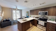 This single-floor residence offers a thoughtfully designed open-concept layout that maximizes both functionality and comfort. The kitchen serves as the heart of the home, showcasing rich dark wood cabinetry paired with crisp white countertops that create a striking contrast. Stainless steel appliances, including a range, built-in microwave, and dishwasher, lend a polished, contemporary feel. The kitchen peninsula features a double undermount sink and provides additional seating and prep space, making it ideal for both everyday use and entertaining. Warm hardwood-style flooring flows throughout, tying the spaces together cohesively. The open layout allows the kitchen to connect effortlessly to the dining and living areas, where natural light pours in through large windows dressed with sheer curtains. A ceiling fan adds both style and practicality to the living space. Recessed lighting throughout ensures the interior remains bright and welcoming at any time of day. The overall ambiance is modern and inviting, with a neutral palette accented by warm wood tones that create a comfortable and stylish living environment.