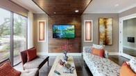 This conference room offers a sophisticated and inviting atmosphere, blending contemporary design elements with warm, natural materials. The centerpiece of the space is a rich, wood-planked accent wall that extends to the ceiling, providing a striking backdrop for a wall-mounted flat-screen display — ideal for presentations and collaborative meetings. Matching wood ceiling panels add continuity and warmth, lending the room an upscale, cohesive aesthetic.

Large floor-to-ceiling windows flood the space with abundant natural light and offer pleasant views of a landscaped garden area, creating a refreshing and open ambiance. Dark, large-format tile flooring runs throughout, grounding the room with a polished, modern finish.

Comfortable seating — including a patterned sofa and tufted accent chairs with coral accent pillows — is thoughtfully arranged to encourage both formal and informal interaction. A light wood console dresser with decorative lamps and metallic wall art adds a refined, curated touch to the entryway area, while an exposed brick wall element and glass partitions contribute architectural character and visual interest.

Recessed lighting and framed wall art with warm tones complete the refined interior, making this conference room a functional yet aesthetically compelling space well-suited for meetings, gatherings, or collaborative work sessions.