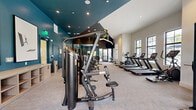 Located at 2085 S Congress Ave in Palm Springs, FL, this professionally designed fitness center spans a generously sized single floor divided into three dedicated exercise zones. The space features a striking contemporary design, with bold teal accent walls contrasting against crisp white surfaces and dark ceiling panels, creating a dynamic and energizing atmosphere throughout.

The cardio zone is lined with large, grid-style windows that flood the space with natural light and offer views of the surrounding palm-tree-lined exterior. A row of premium treadmills, ellipticals, and stationary bikes — all from the Hoist brand — are positioned to take full advantage of the natural light and outdoor views. Recessed lighting throughout ensures the space remains bright and welcoming at all hours.

The strength training zone is equally well-appointed, featuring an extensive selection of Hoist cable machines, weight benches, and functional training equipment. Full-length mirrors amplify the sense of space, while the neutral-toned carpet flooring provides comfort underfoot. A third area offers additional multi-use exercise space.

Thoughtful amenities include built-in wood cabinetry for personal storage, a water station with a tiled surround, and framed fitness-themed artwork that adds a refined, boutique-gym aesthetic. The high, vaulted ceilings and open floor plan contribute to an airy, spacious feel befitting a premium facility.