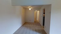 This single-floor residence offers a straightforward and functional layout designed for comfortable everyday living. The main living area is finished with wall-to-wall carpeting in a neutral beige tone, creating a warm and cohesive feel throughout the space. A ceiling fan with integrated lighting is centrally mounted, providing both air circulation and ambient illumination to the room.

The living area opens through an arched doorway into a hallway that connects the various areas of the home. A floor vent is visible along the baseboard, indicating a built-in HVAC system for year-round climate control. The walls are painted in a clean, neutral white that reflects light and contributes to a bright, open atmosphere.

Visible through the adjacent opening is a kitchen area featuring dark cabinetry, offering a contrast to the lighter tones found throughout the rest of the space. The layout suggests a practical flow between the living, hallway, and kitchen areas, making the home easy to navigate.

Overall, the space presents a clean, move-in-ready environment with a neutral palette and functional design elements. The carpeted flooring, ceiling fan, and connected layout make it a practical and livable single-floor home.