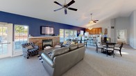 This single-floor clubhouse is designed to accommodate a variety of community activities and social gatherings, offering an open, well-appointed layout with distinct functional zones flowing seamlessly into one another.

The centerpiece of the space is a generously sized great room with soaring vaulted ceilings and multiple ceiling fans that enhance the airy atmosphere. A bold navy accent wall anchors the living area, where leather seating is arranged around a striking stone fireplace topped with a wall-mounted television — creating an inviting focal point for relaxation and entertainment.

Adjacent to the seating area, a billiards table provides recreational fun, while multiple dining tables and chair sets offer ample space for communal meals or casual gatherings. Large windows throughout the room flood the interior with natural light and provide pleasant outdoor views.

The kitchen area features warm wood cabinetry, a breakfast bar with seating, and standard appliances including a refrigerator, making it well-suited for catering events or community use. Wainscoting details along the lower walls add a classic, polished character throughout the common areas.

A separate, sunlit room — visible through a pass-through window — provides additional flexible space, suitable for meetings or office use. The carpeted flooring throughout contributes to a comfortable and inviting ambiance, rounding out this thoughtfully designed community clubhouse.