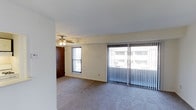 This single-floor space centers around a spacious, open living area finished with wall-to-wall carpeting that lends a comfortable, understated feel underfoot. Large sliding glass doors dominate one wall, fitted with vertical blinds that allow for flexible light control and provide views of the surrounding exterior. An additional window further enhances the natural light flowing into the room, keeping the space feeling bright and airy throughout the day. A ceiling fan with an integrated light fixture is centrally positioned, offering both ventilation and supplemental lighting. The walls are finished in a clean, neutral off-white tone that complements the light palette of the room. A solid wood door is visible toward the rear of the living area, suggesting access to additional spaces within the unit. The overall layout is open and straightforward, with a simple, functional design that provides a versatile canvas for furnishing and personalizing. The single-floor configuration ensures easy accessibility and a seamless flow throughout the space.