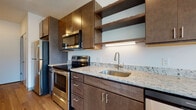 This well-appointed single-floor space showcases a thoughtfully designed interior with a consistent modern aesthetic throughout. The kitchen serves as a central highlight, featuring rich dark wood cabinetry paired with light granite countertops that provide an elegant contrast. A stainless steel appliance suite—including a refrigerator, electric range, over-the-range microwave, and dishwasher—offers full functionality. Open shelving above the sink adds a contemporary touch, while under-cabinet lighting enhances visibility and ambiance. A peninsula with a matching granite surface extends from the kitchen, creating a natural transition into the open living area and offering additional workspace or casual seating.

The living area is defined by warm, light-toned hardwood flooring that runs consistently throughout the space, contributing to a cohesive and inviting atmosphere. Clean white walls and recessed lighting keep the interior feeling bright and open.

The bedroom is generously proportioned, bathed in natural light through a large picture window that frames views of the surrounding neighborhood and mature trees, creating a pleasant indoor-outdoor connection.

The bathroom features a clean, minimalist walk-in shower with white surround panels, a built-in brushed nickel fixture, and coordinating towel bars, delivering a streamlined and functional finish. Overall, the space presents a polished, contemporary living environment.