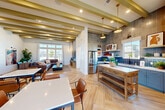 Located at 448 Denton Ave in Vacaville, CA, this single-story community clubhouse offers a well-appointed gathering space designed with modern, contemporary finishes throughout. The interior showcases striking exposed beam ceilings finished in an earthy olive tone, herringbone-patterned wood-look flooring, and crisp white walls that create a bright, welcoming atmosphere.

The open-plan layout seamlessly connects the kitchen, dining, and lounge areas. The kitchen is a standout feature, appointed with bold blue tile backsplash, open shelving, stainless steel appliances, and a rustic wood island. Multiple dining tables with leather chairs provide ample seating, while large windows and glass sliding doors bathe the interior in abundant natural light and offer views of the surrounding neighborhood.

The lounge area is anchored by a comfortable sectional sofa, decorative pendant lighting, and a feature wall with a mounted display, creating a relaxed social environment. A smaller breakout seating nook with floor-to-ceiling glass doors leads directly to the outdoor patio.

Three well-maintained bathrooms with tiled walls and commercial-grade fixtures serve the space. Outdoors, a generously sized swimming pool is surrounded by a spacious sun deck lined with lounge chairs and shaded by blue umbrellas, all enclosed within a sleek black metal fence with manicured landscaping.